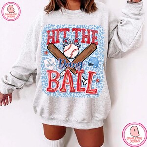 May include: A light gray sweatshirt with a baseball-themed graphic. The graphic features a baseball, crossed bats, and the words "Hit the Dang Ball" in red. A blue leopard print background completes the design.