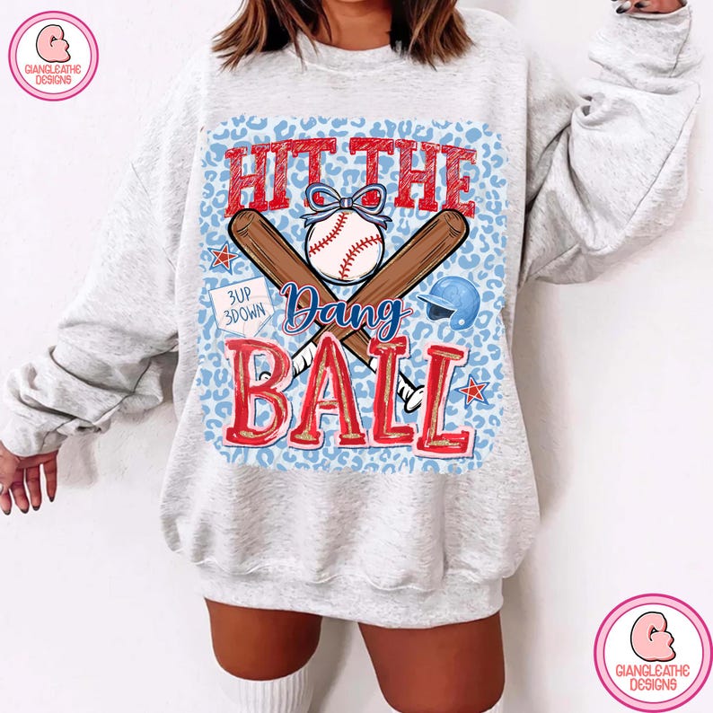 May include: A light gray sweatshirt with a baseball-themed graphic. The graphic features a baseball, crossed bats, and the words "Hit the Dang Ball" in red. A blue leopard print background completes the design.