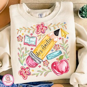 May include: Cream-colored sweatshirt featuring a colorful teacher-themed graphic. The design includes a large yellow pencil with the name "Mrs. Amelia", a red apple, an open book, and floral elements. A paper airplane and ruler are also included.