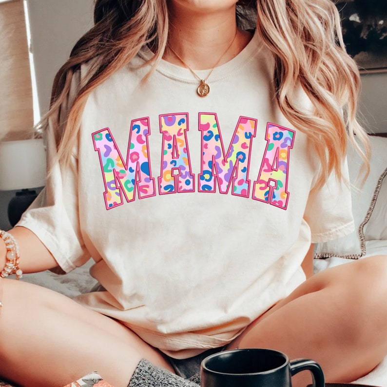 May include: A white t-shirt with the word "MAMA" printed in large, pink outlined letters. The letters are filled with a colorful leopard print pattern.