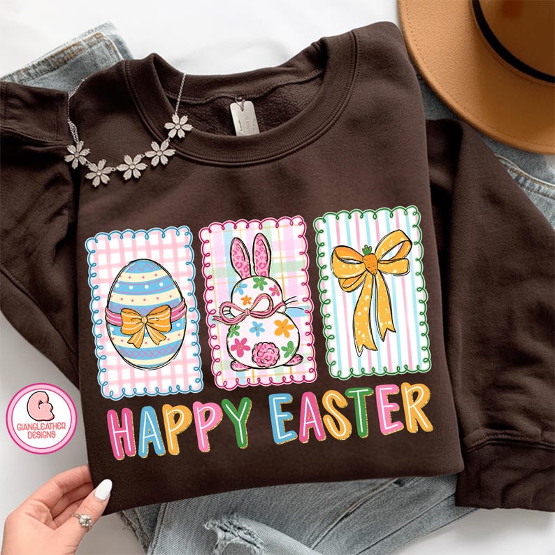 May include: A brown sweatshirt featuring three Easter-themed designs. The designs include a decorated egg, a bunny, and a bow, all within decorative frames. The words "HAPPY EASTER" are printed in colorful letters below the designs.