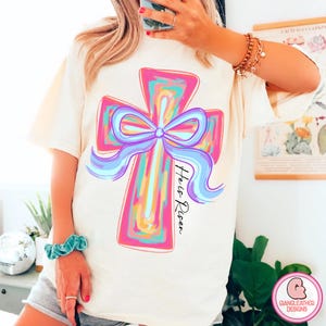 May include: A white t-shirt with a colorful painted cross and a purple bow. The text "He is Risen" is written on the shirt.