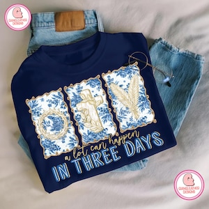 Könnte beinhalten: Marineblaues T-Shirt mit floralem und religiösem Design. Das Shirt zeigt drei gerahmte Bilder mit einem Kreuz, einer Dornenkrone und einer Weizenähre. Der Text "a lot can happen IN THREE DAYS" ist in Gold und Blau gedruckt.