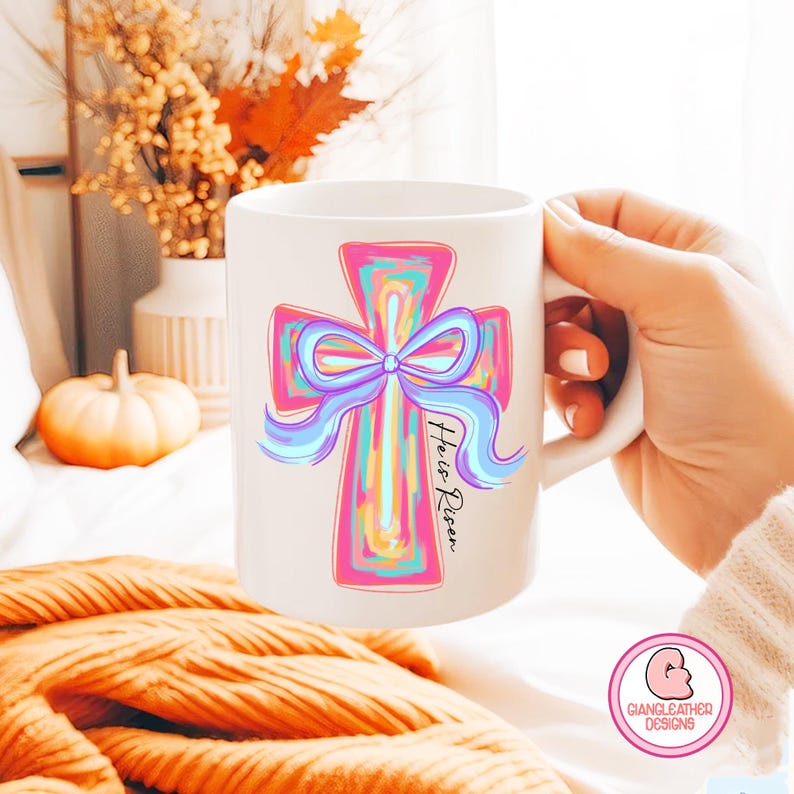 May include: White ceramic mug with a pink, blue, and yellow cross with a blue bow and the text "He is Risen".