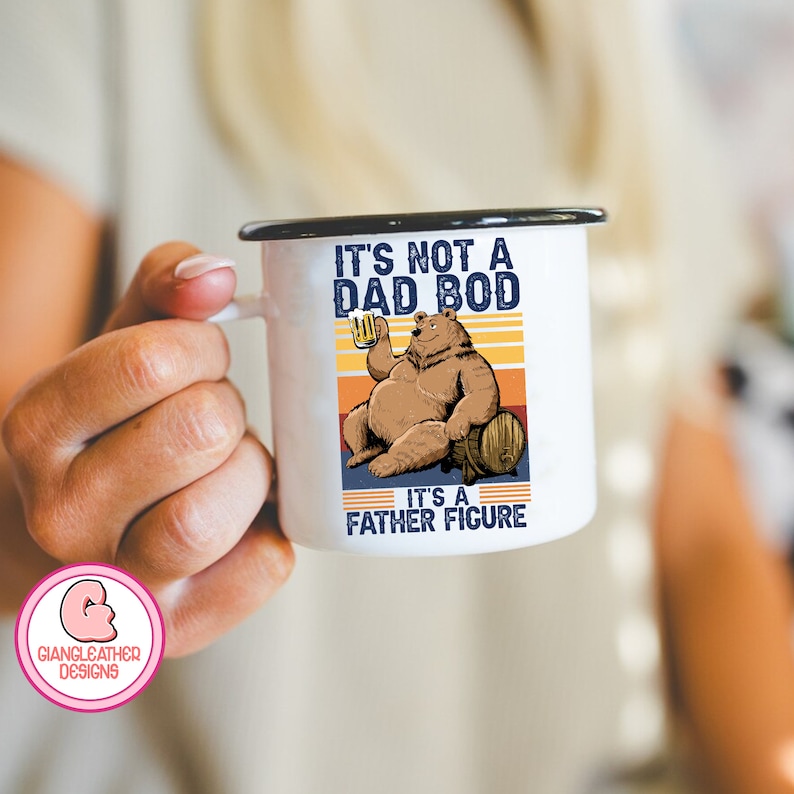 May include: White enamel mug with a black rim and handle. The mug features a cartoon illustration of a brown bear sitting on a barrel, holding a beer. The text on the mug reads "It's not a dad bod, it's a father figure."