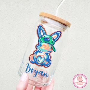 May include: A clear glass reusable water bottle with a bamboo lid and a straw. The bottle has a colorful cartoon bunny wearing a green hat and the name "Bryan" printed on the side.