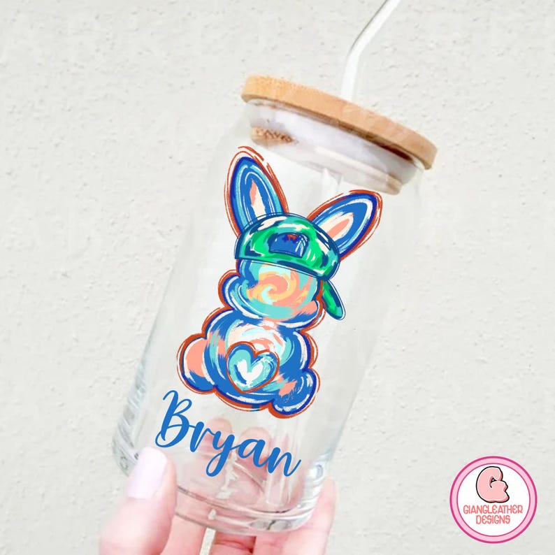May include: A clear glass reusable water bottle with a bamboo lid and a straw. The bottle has a colorful cartoon bunny wearing a green hat and the name "Bryan" printed on the side.