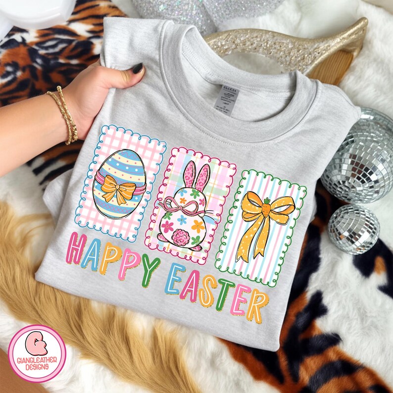 May include: A light gray t-shirt featuring three Easter-themed designs. The designs include a decorated egg, a bunny, and a bow, all within decorative frames. The words "HAPPY EASTER" are printed in colorful letters below the designs.