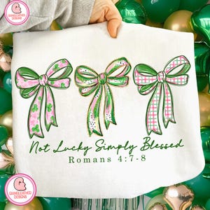 Puede incluir: Diseño blanco con temática del Día de San Patricio con tres lazos verdes y rosas con diferentes patrones. El texto "Not Lucky Simply Blessed" y "Romans 4:7-8" está impreso en verde debajo de los lazos. El diseño es sostenido por una persona.