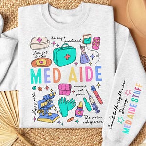 May include: A gray sweatshirt with a colorful graphic design featuring the words "MED AIDE" and various medical symbols, including a first aid kit, pills, a stethoscope, and a syringe. The text "Can't talk right now Doing MED AIDE STUFF" is printed on the sleeve.