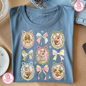 K&ouml;nnte beinhalten: Hellblaues T-Shirt mit einem Design von Osterhasen in verzierten Rahmen, umgeben von Schleifen und floralen Akzenten. Die Hasen halten Ostereier, Blumen und Karotten. Das T-Shirt hat einen runden Halsausschnitt.