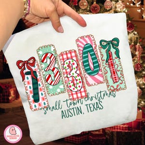 Könnte beinhalten: Weißes T-Shirt mit festlichem Weihnachtsdesign. Das Design zeigt die Zahlen "1301" in einer farbenfrohen, weihnachtlichen Schriftart mit Weihnachtsbaum-, Zuckerstangen- und Schleifenakzenten. Der Text "small town christmas AUSTIN, TEXAS" steht darunter.