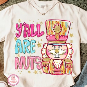 Op de afbeelding: Wit T-shirt met een kleurrijke afbeelding van een notenkraker en de tekst "Y'all Are Nuts" in roze en blauw. De notenkraker is roze, goud en blauw met een grote snor en een hoge hoed. Gouden sterren zijn verspreid rond het ontwerp.