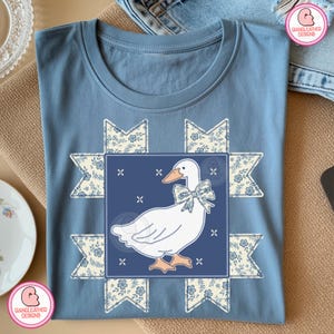 Peut inclure: Un t-shirt bleu clair avec un motif d'oie blanche orné d'un nœud papillon bleu, encadré d'éléments à motifs floraux. Le t-shirt est plié avec soin, mettant en valeur le motif central. Le design comprend le texte "GIANGLEATHER DESIGNS".