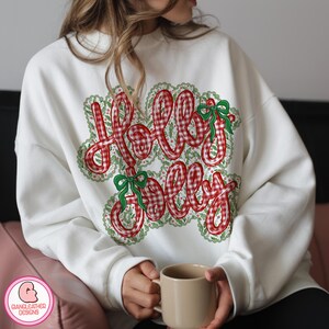 Puede incluir: Sudadera blanca con las palabras "Holly Jolly" en estampado de cuadros rojos. El texto est&aacute; delineado con un dise&ntilde;o floral verde y blanco y acentuado con lazos verdes. Una prenda con tem&aacute;tica navide&ntilde;a.