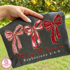 May include: Dark gray t-shirt featuring three red bows with gold trim. The bows have a mix of solid and gingham patterns. The text "Oh, how He loves us" and "Ephesians 2:4-8" are printed in red. A logo is in the lower left corner.