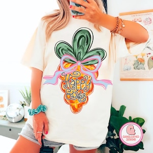 May include: White t-shirt with a graphic of a cartoon carrot with a green top and a pink bow. The text "Happy Easter" is written on the carrot.
