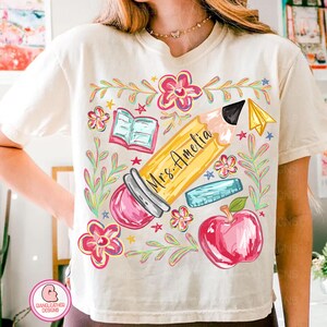 May include: Cream-colored t-shirt featuring a colorful teacher-themed graphic. The design includes a large yellow pencil with "Mrs. Amelia" written on it, surrounded by flowers, a book, a ruler, a red apple, and a paper airplane.