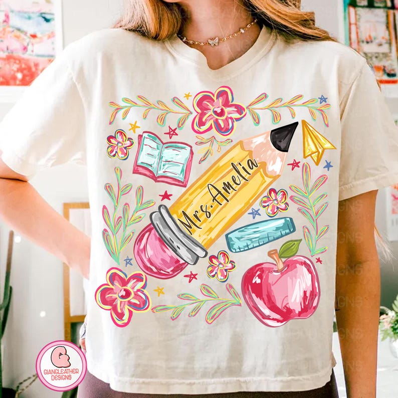 May include: Cream-colored t-shirt featuring a colorful teacher-themed graphic. The design includes a large yellow pencil with "Mrs. Amelia" written on it, surrounded by flowers, a book, a ruler, a red apple, and a paper airplane.