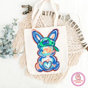 May include: A white tote bag with a blue and green bunny wearing a green baseball cap. The bunny has a heart on its chest and is painted in a colorful abstract style.