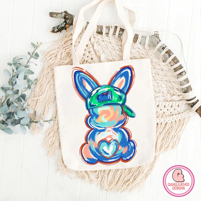 May include: A white tote bag with a blue and green bunny wearing a green baseball cap. The bunny has a heart on its chest and is painted in a colorful abstract style.
