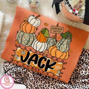 May include: Orange long-sleeve shirt with a pumpkin patch design. The design features stacks of pumpkins in orange, white, and camouflage green, with the name "JACK" in large black letters. A sign reads "Pumpkin Patch".