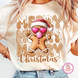 May include: White t-shirt with a gingerbread man wearing pink heart-shaped sunglasses and a red Santa hat. The gingerbread man is holding up a peace sign. The text "Merry Merry Christmas" is printed on the shirt in brown.