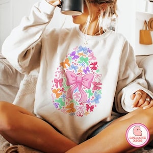 May include: A white sweatshirt with a colorful design of bows and hearts in the shape of an egg. The design is centered on the front of the sweatshirt.