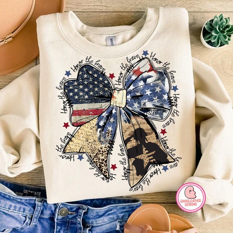 May include: A beige sweatshirt with a large bow made of an American flag and a leopard print fabric. The bow is surrounded by the words "Honor the brave" repeated multiple times.