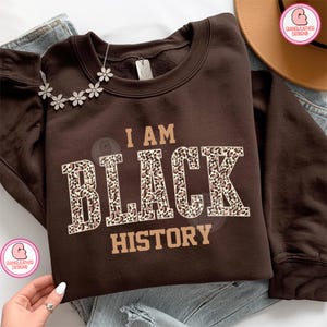 Puede incluir: Una sudadera marrón con el texto "I AM BLACK HISTORY" en beige y estampado de leopardo. Un collar de flores plateado descansa sobre el hombro. La sudadera está doblada sobre unos vaqueros azules.