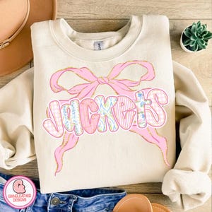 May include: Cream-colored sweatshirt with the word "JACKETS" in a pink, patterned font, accented by a pink bow with gold glitter trim. The sweatshirt is displayed on a wooden surface with a hat and jeans.