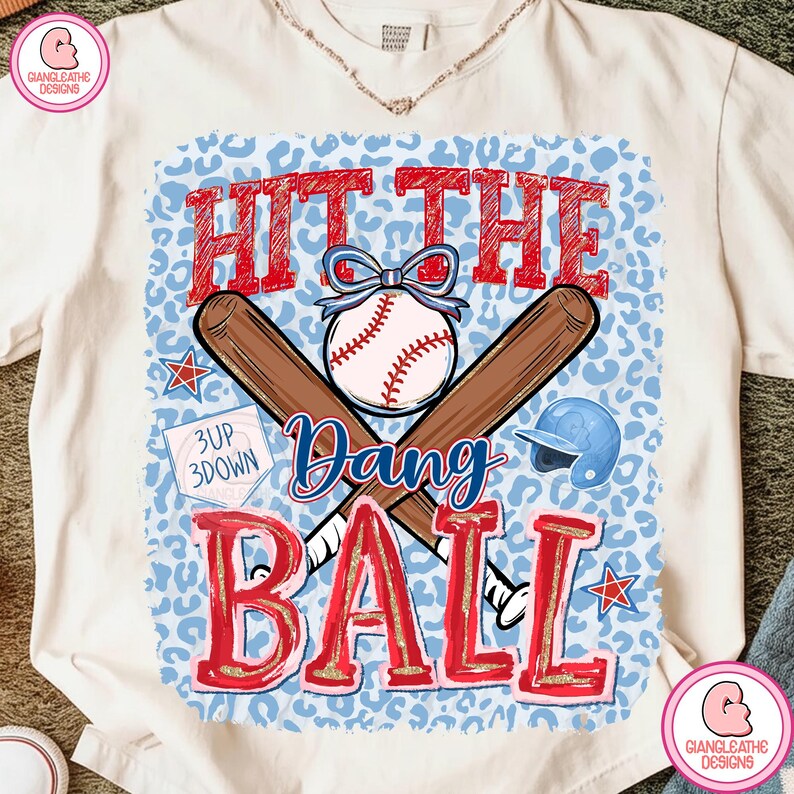 May include: Cream-colored t-shirt with a baseball-themed graphic. The design features the words "Hit the Dang Ball" in red, a baseball, crossed bats, a helmet, and stars. The background has a blue leopard print.