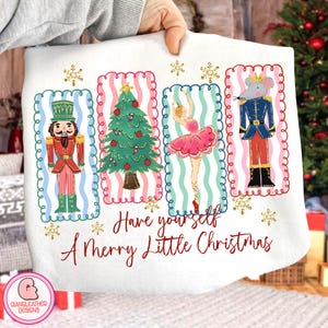 May include: White Christmas sweatshirt featuring a nutcracker, Christmas tree, ballerina, and mouse soldier illustrations. The text "Have yourself A Merry Little Christmas" is written in red script. Gold snowflakes decorate the design. Festive and seasonal.