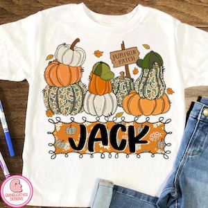 May include: White t-shirt featuring a pumpkin patch design. The design includes stacked pumpkins in orange, white, and camouflage patterns, with a sign that reads "Pumpkin Patch." The name "JACK" is displayed in a decorative frame.