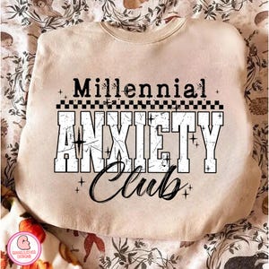 May include: A cream-coloured sweatshirt featuring the text "Millennial Anxiety Club" in black and white. The words are in a bold, distressed font, with a checkered pattern above "Anxiety". The sweatshirt is shown on a floral patterned fabric.