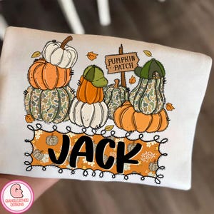 May include: White long-sleeve shirt featuring a stacked pumpkin design with orange, white, and camouflage pumpkins. The shirt includes the text "PUMPKIN PATCH" and the name "JACK" in a decorative font, perfect for fall.