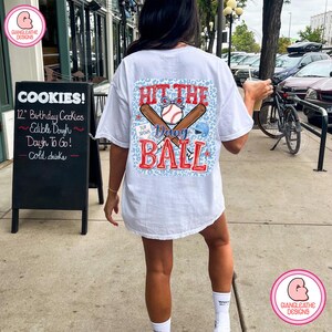 May include: White t-shirt with a baseball-themed graphic on the back. The design includes a baseball, bats, and the text "Hit the Dang Ball" in red and blue. The shirt is worn by a person standing on a sidewalk.