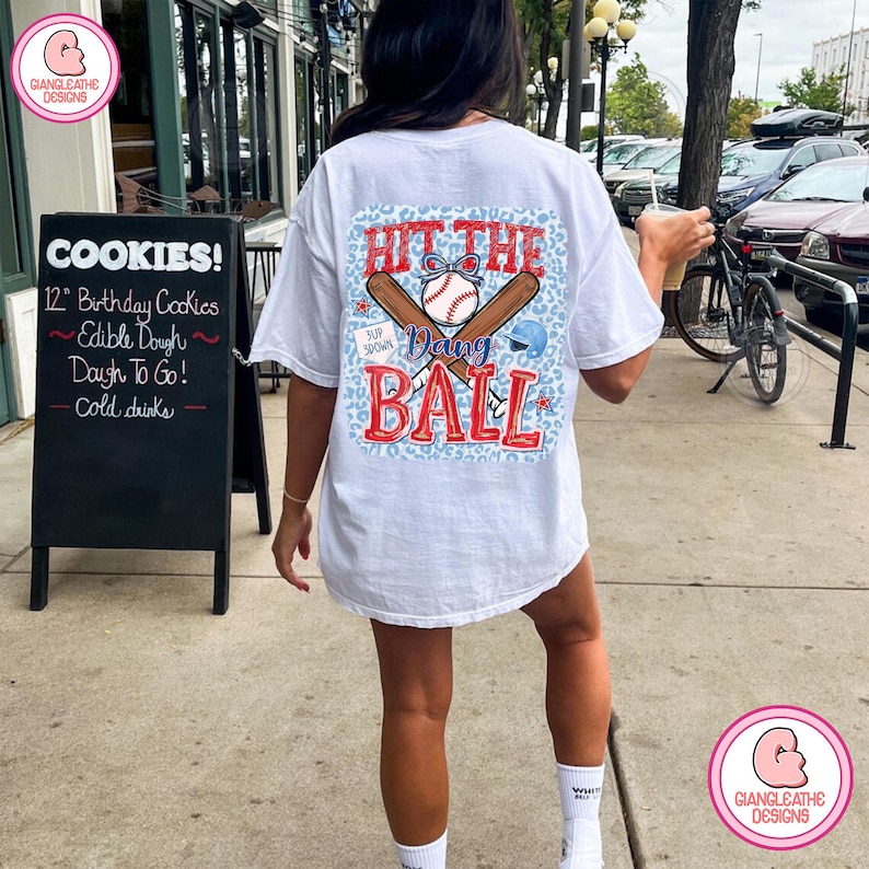 May include: White t-shirt with a baseball-themed graphic on the back. The design includes a baseball, bats, and the text "Hit the Dang Ball" in red and blue. The shirt is worn by a person standing on a sidewalk.