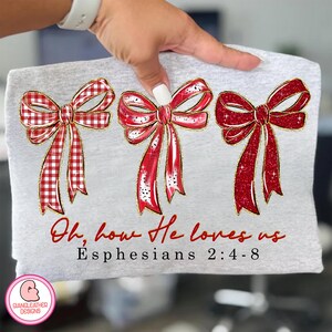 May include: A light gray t-shirt featuring three red bows with gold trim. The bows have a mix of solid and plaid patterns. The text "Oh, how He loves us" and "Ephesians 2:4-8" are printed in red script.