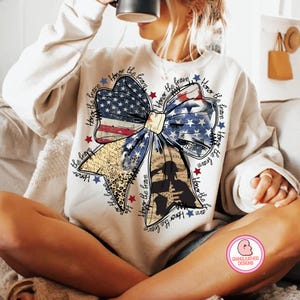 May include: A white sweatshirt with a large bow made of an American flag print. The bow is surrounded by the text "Honor the brave" repeated multiple times.