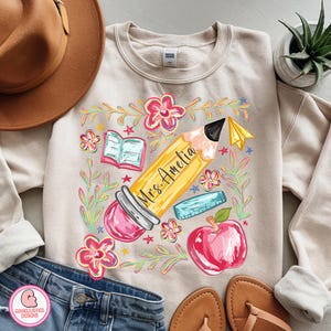 May include: Beige sweatshirt with a colorful graphic featuring a large yellow pencil with the text "Mrs. Amelia", surrounded by flowers, an apple, a book, and a ruler. The design is in a whimsical, cartoon style.