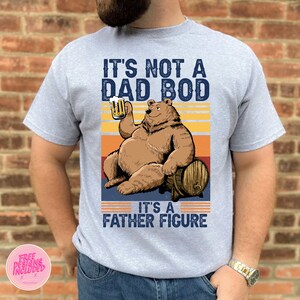 May include: A gray t-shirt with a vintage-style graphic of a bear holding a beer and sitting on a barrel. The text on the shirt reads "It's not a dad bod, it's a father figure."
