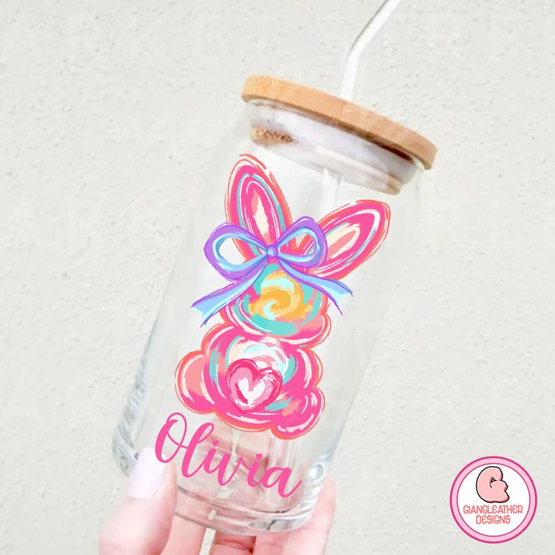 May include: A clear glass jar with a bamboo lid and a straw. The jar has a colorful painted Easter bunny design with a pink bow and the name "Olivia" written on the front.