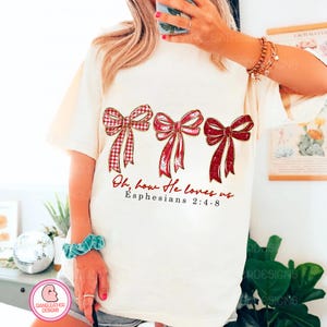 May include: Cream-colored t-shirt featuring three red bows with gold accents. The bows have different patterns, including plaid and solid designs. The text "Oh, how He loves us Ephesians 2:4-8" is printed below the bows. The shirt is a casual style.