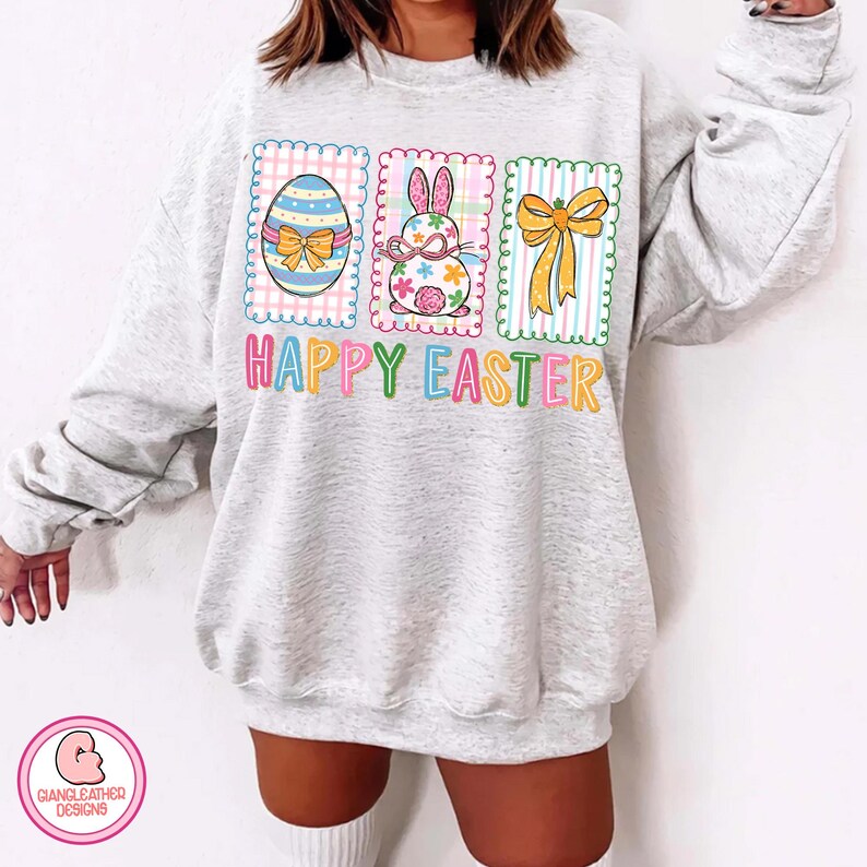 May include: A light gray sweatshirt with a colorful Easter graphic. The graphic includes an Easter egg, a bunny, and a bow, with the words "HAPPY EASTER" in bright letters. The design is framed by a scalloped border.