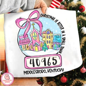 May include: White pillow with a snow globe design featuring a pink church, buildings, and a Christmas tree. The text reads "Christmas is better in a small town" and "Middlesboro, Kentucky 40965". A pink bow is at the top.