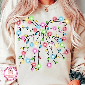 May include: A white sweatshirt featuring a black outline of a bow made from Christmas lights. The lights are in various colours, including red, green, blue, yellow, and pink.