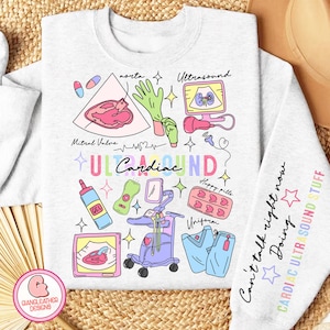 May include: A light gray sweatshirt with a colorful graphic design of a heart, ultrasound machine, and other medical equipment. The text "ULTRA Cardiac SOUND" is written in rainbow colors. The text "Can't talk right now Doing CARDIAC ULTRASOUND STUFF" is written on the sleeve.