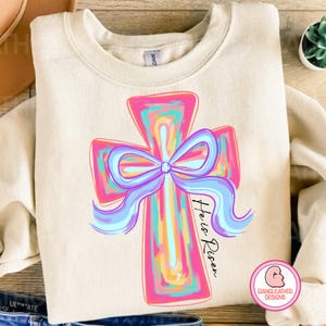 May include: A white sweatshirt with a colorful cross and a blue bow. The text "He is Risen" is written on the sweatshirt.