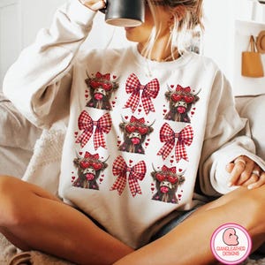 Può includere: Felpa bianca con un fiocco a quadri rosso e bianco e una mucca delle Highlands con occhiali a forma di cuore e un lecca-lecca rosso. Il design della mucca è ripetuto otto volte sulla felpa.